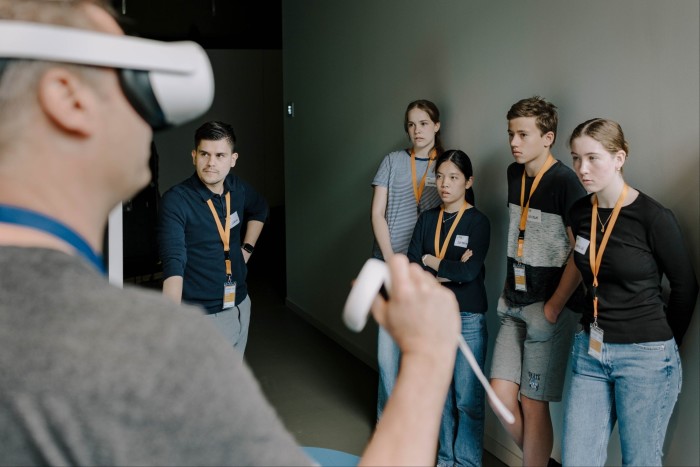 A group of young adults look intently at a man wearing a virtual reality headset