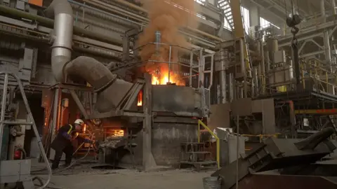 BBC News / Kevin Church Flames emerge from the top of the arc furnace as material is added to the molten steel