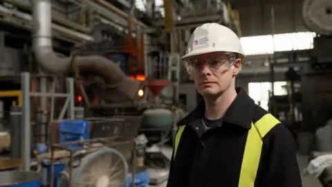 BBC News / Kevin Church Image of scientist Dr Cyrille Dunant standing in front of the electric arc furnace at the Materials Processing Laboratory in Middlesborough.  The oven glows red hot.