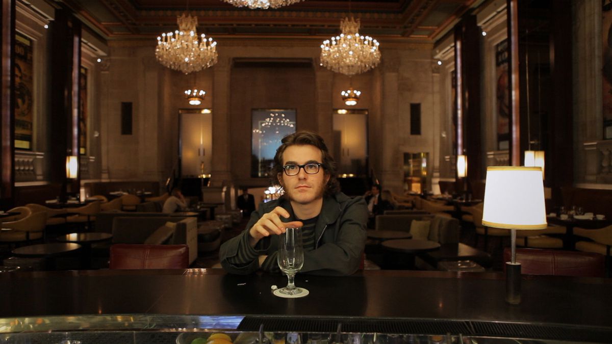 Phil Fish, wearing hipster glasses and mutton chops, has a drink in a chic hotel bar