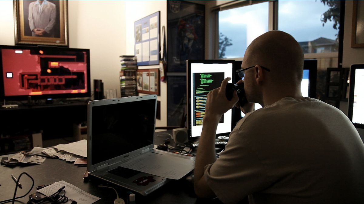 Tommy Refenes sits at his desk, surrounded by computer screens, playing Super Meat Boy on a TV in the background