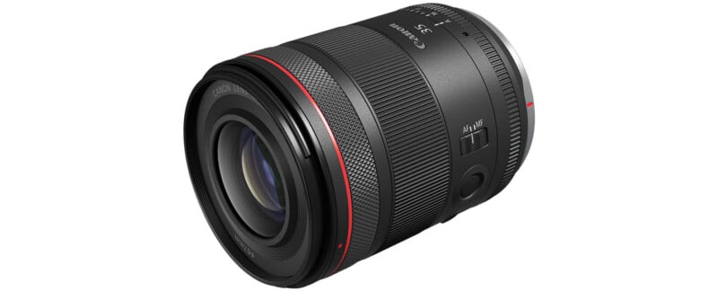 A black Canon RF 35mm f/1.8 IS Macro STM lens is shown against a white background.  The lens is equipped with a wide focus ring, a control ring and an image stabilization switch.  It has a red ring near the front element, indicating its L-series status.