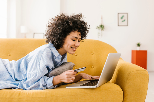 Smiling woman with credit card using laptop lying on sofa in living room at home