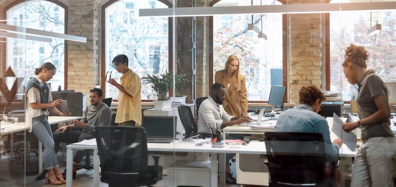 A modern office with brick walls and large windows.  Several people work at a desk with computers, while others stand and talk.  There are factories and office equipment everywhere, creating a collaborative and busy work environment.