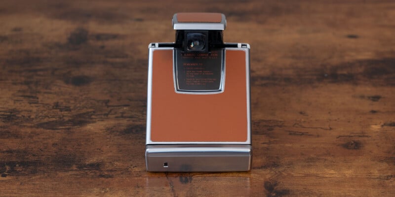 A vintage Polaroid SX-70 land camera with a brown leather exterior and a silver metal frame is placed on a wooden surface.  The camera is in a closed position, which shows off its rectangular shape and sleek design.