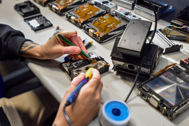 You see a person's hands working at a table full of electronic components and tools.  One hand uses a soldering iron while the other holds a green tool.  Various dismantled devices and components, including hard drives, are scattered across the workspace.