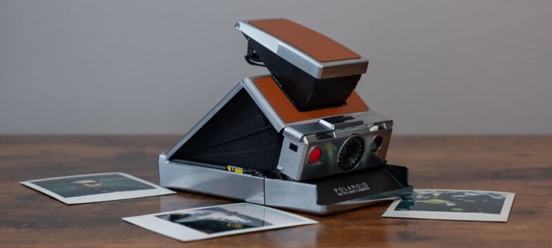 A vintage Polaroid SX-70 land camera with a brown leather exterior sits on a wooden surface.  The camera is partially extended.  Two instant photos are scattered in the foreground, partially visible.  The background is a plain gray wall.