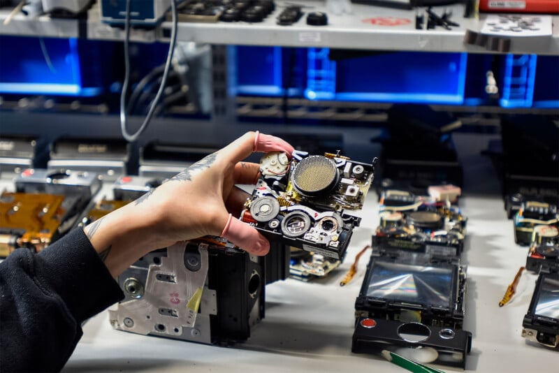 A person holds a disassembled mechanical part, possibly from a vintage electronic device, in a workshop.  The workspace is cluttered with additional disassembled components and tools, with shelves in the background illuminated by blue lights.