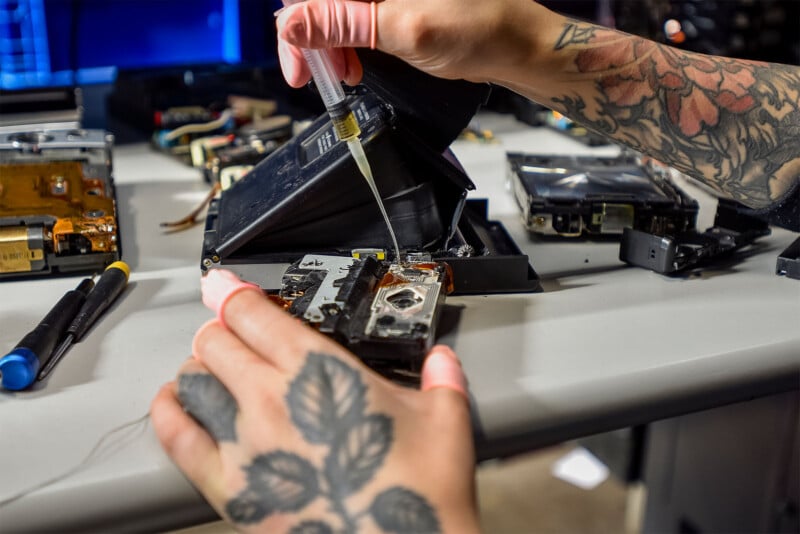 A person with tattooed hands and pink gloves repairs electronic components at a workstation.  They use a syringe to apply liquid to a part, while various tools and electronic devices lie on the table.