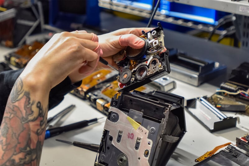 A person with tattooed arms disassembles a camera, while various small tools and electronic components are scattered on the workbench.  The background consists of shelves with more electronic equipment and a computer screen.