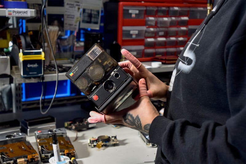 A person holding a vintage Polaroid camera in a workshop.  Various electronic components, tools and storage drawers can be seen in the background, indicating a repair or restoration environment.  The person's hand tattoo and a partial view of the sweatshirt are visible.