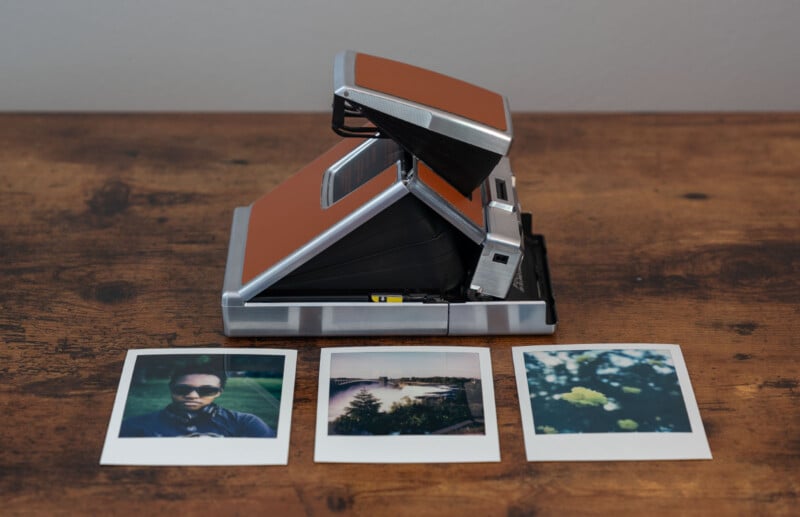 A vintage instant camera with a brown exterior stands on a wooden surface.  In front of it are three Polaroid photos: one of a person wearing sunglasses, one of a scenic view with a body of water, and one of yellow flowers in the foreground.