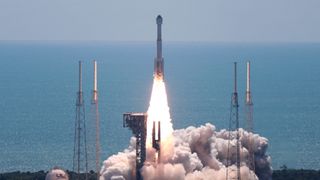 a white rocket is launched above a plume of fire and smoke on a sunny day