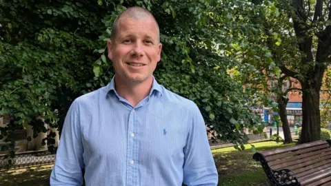 Ben Perks Mortgage Advisor Ben Perks is smiling, wearing a light blue shirt and standing in front of a bench and trees