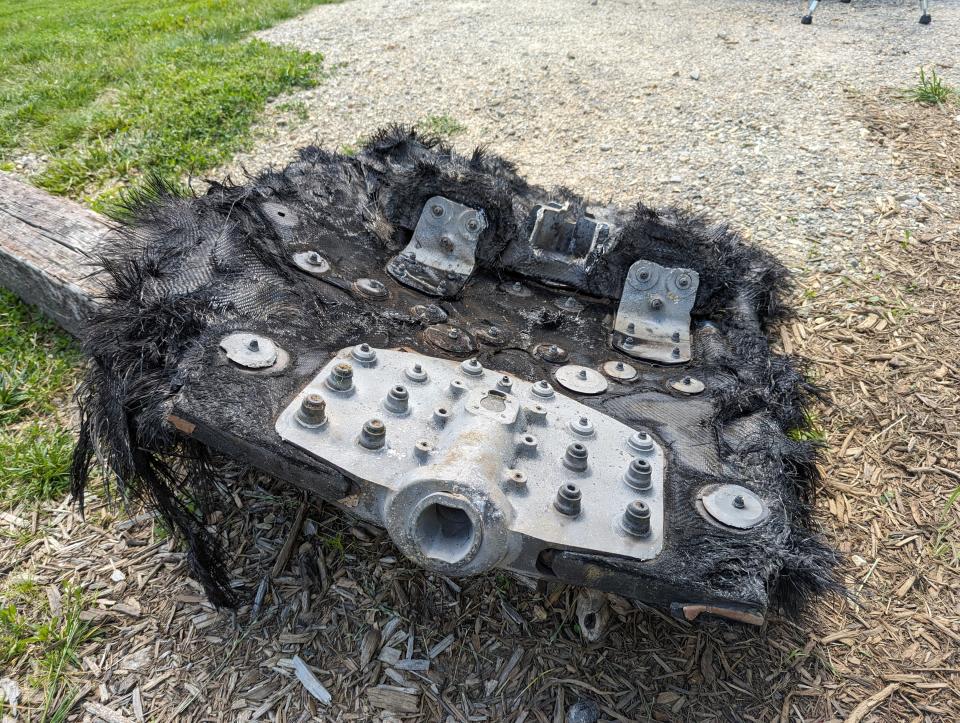 a large black piece of fiberglass covered in metal bolts and plates lies on the ground next to a path leading to a forest.  Mountains can be seen rolling in the distance