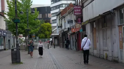 A few women walk down a street in Swindon, with a man in a white hoodie and a stool on the right, in front of a boarded up shop, a boarded up shop in the foreground