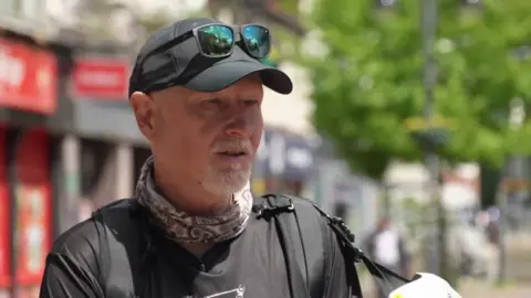 Gary Huett wears a black baseball cap with sunglasses on top, a black T-shirt and a patterned scarf around his neck