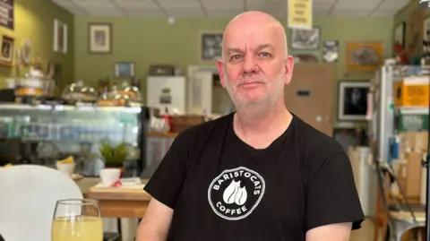 Marcus Kittridge sits in his cafe wearing a black T-shirt that says Baristocats Cafe on it, with a glass of orange juice on the table in front of him