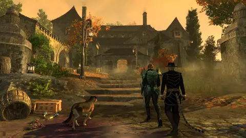 Bethesda Screenshot from Elder Scrolls Online shows two humanoid characters – one human and one with a tail – approaching a large mansion-style building at sunset.  There is some fog hanging along the approach, which is littered with barrels and similar objects, and a large feline companion walks alongside them.