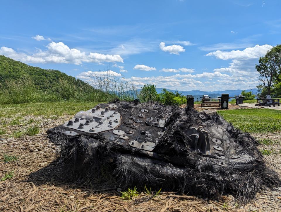 a large black piece of fiberglass covered in metal bolts and plates lies on the ground next to a path leading to a forest.  Mountains can be seen rolling in the distance