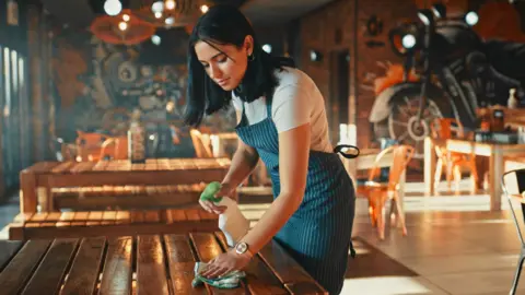 Getty Images A young woman with dark hair works in a cafe and cleans a table