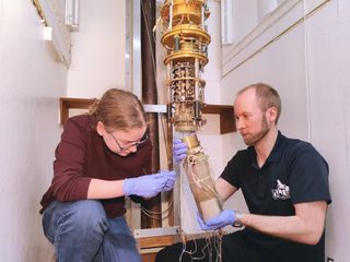 A white room with two crouched people operating a complicated gold machine