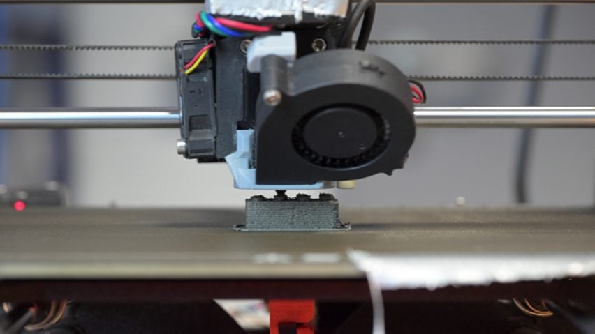 Close-up photo of a 3D printer producing a small, gray, Lego-like block