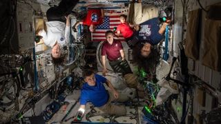 several astronauts in a dark module of the space station with their faces and bodies illuminated by camera lights. an American flag can be seen in the background