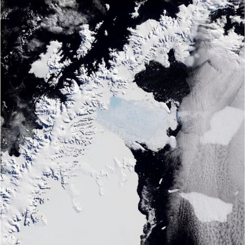 A satellite image of a largely snow-covered landmass with visible dark waters and scattered icebergs. The left side has darker patches of water alternating with white ice formations, while thick clouds dominate the right side of the image.