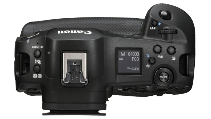 Top view of a Canon digital camera showing buttons, dials and an LCD screen displaying settings including mode "M"ISO 102400 and drive mode. The camera has a textured grip on the right side and a hot shoe mount in the middle.