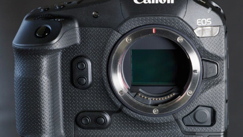 Close-up of a Canon EOS camera body, showing finer details and front panel buttons. The lens has been removed, revealing the sensor in the lens mount. The camera has a textured grip and a sleek black finish.