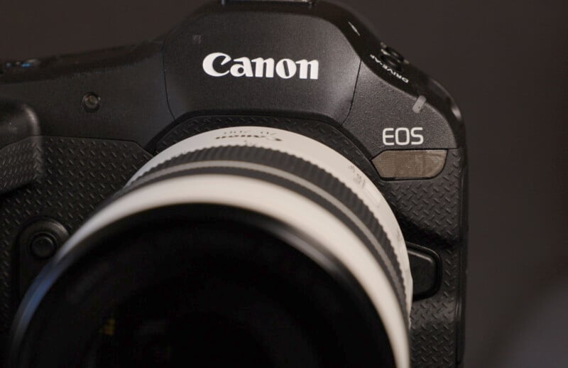 Close-up of a black Canon EOS camera with part of a white lens attached. The word "Canon" is prominently displayed on the top of the camera body and the model designation "EOS" is visible on the right. The background is dark and out of focus.