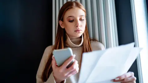 Getty Images Woman standing in front of radiator looking at energy bill with her phone in her other hand