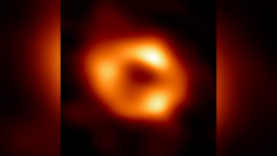 Telescopic image of a black hole, visible as a fuzzy donut of yellow-orange light surrounding a black center