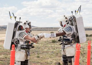 two people in skeletal spacesuits without outer shells walk through the desert under a bright sun