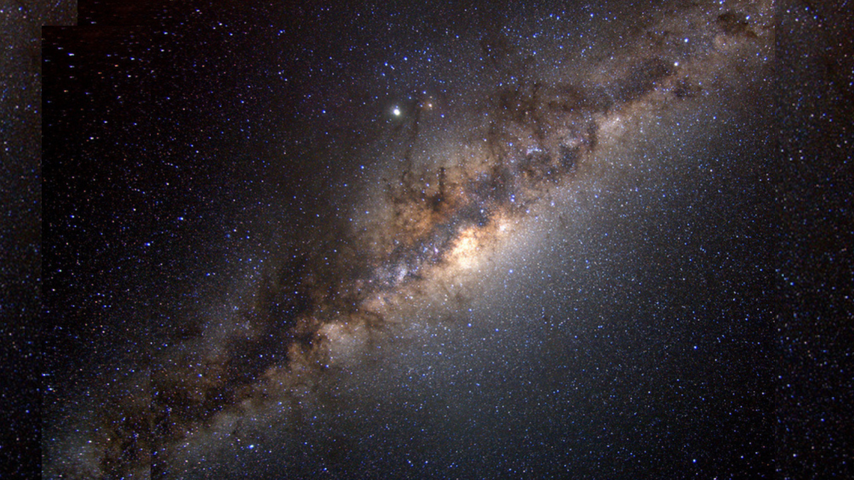 Lots of stars are seen in the image. A central, straight structure goes diagonally from the bottom left to the top right; it has strings of gas, bright sections and other features. It represents the Milky Way