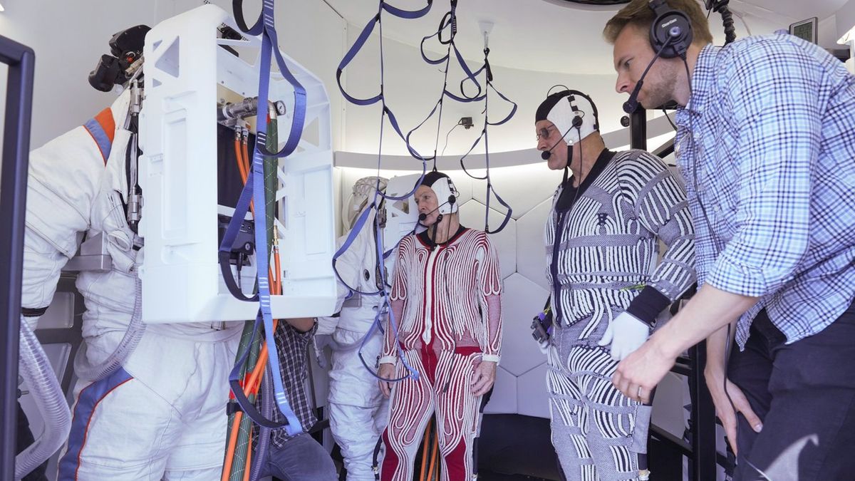 astronauts in white spacesuits stand inside a white airlock