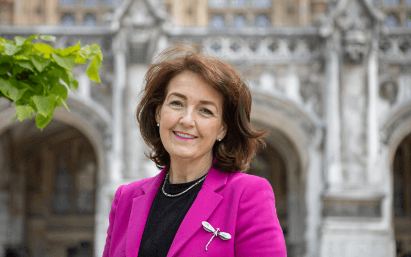 Jo Churchill, head and shoulders, smiling, in a pink jacket, in front of an old building