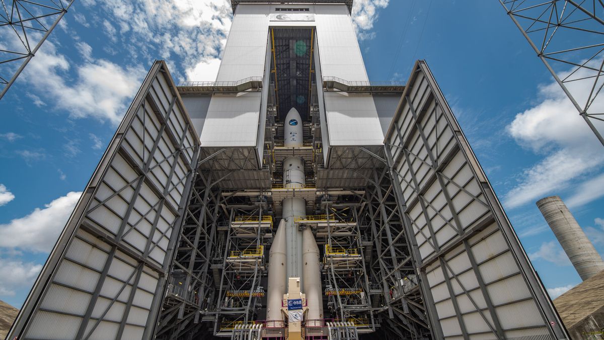 a rocket stands tall in an open assembly hanger before a blue, partly cloudy sky.