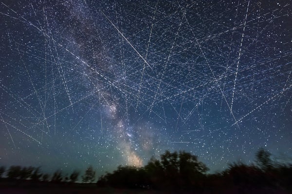 Long exposure composite photography shows light trails from low Earth orbit satellites filling the sky, a tree line is in silhouette at the bottom of the frame