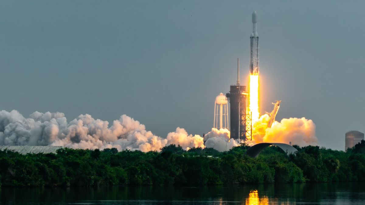 a triple booster rocket blasts off from a launch tower with a blue hazy sky behind