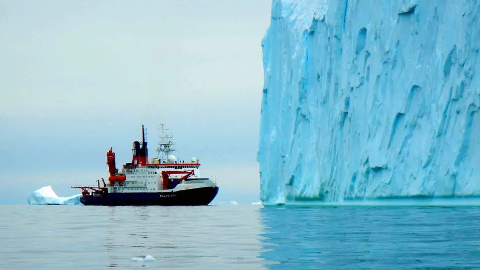 A decent sized boat floats in ocean water next to the vertical incline of an ice cliff.